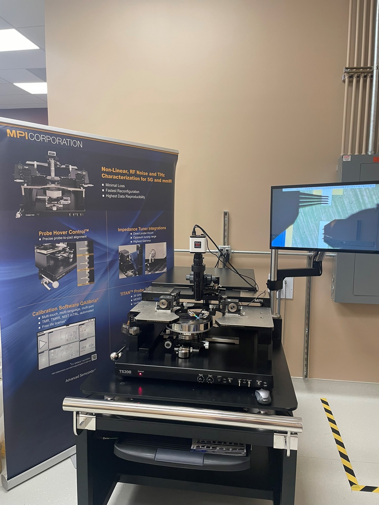 MPI TS200 Manual Probe Station on a black table with a monitor and a promotional banner in a lab setting.