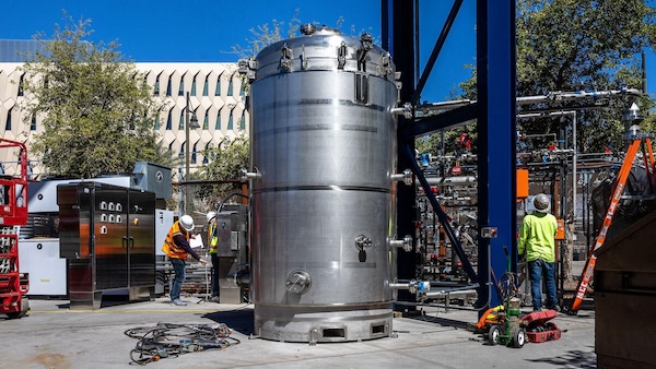 Large stainless steel cylindrical tank, part of the MechanicalTree, in an outdoor industrial setting with workers in high-visibility vests and safety helmets, surrounded by various industrial equipment and a modern building in the background.