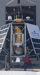 Two people stand in front of the STO-2 telescope, which is flanked by solar panels. The telescope will hang underneath a large helium balloon, which is not inflated in this picture.