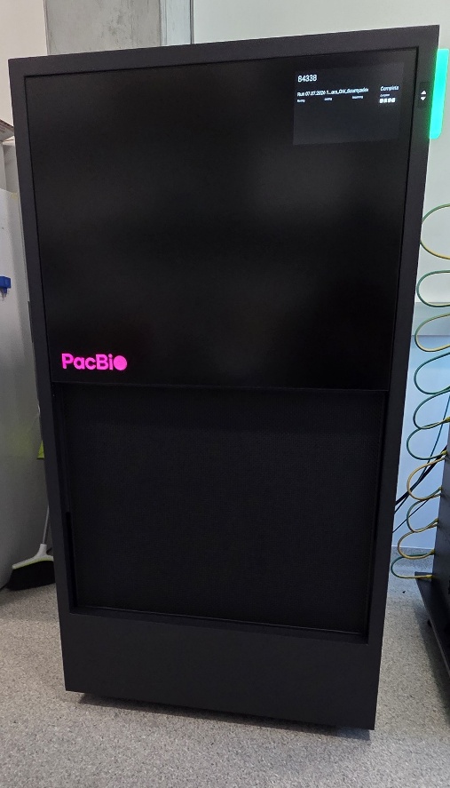 A black PacBio Revio long-read genome sequencer with a pink logo and a small digital screen, featuring a mesh grille on the lower section, set against a laboratory backdrop.